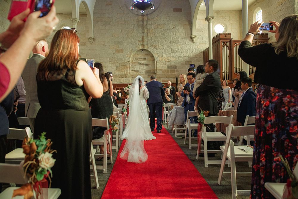 Wedding ceremony in progress with red carpet aisle and white chairs in a church setting.