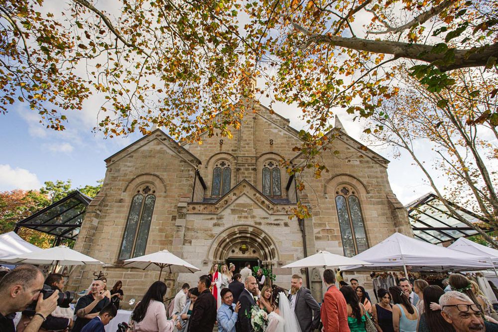 Large group gathers outside historic stone church during autumn wedding celebration.