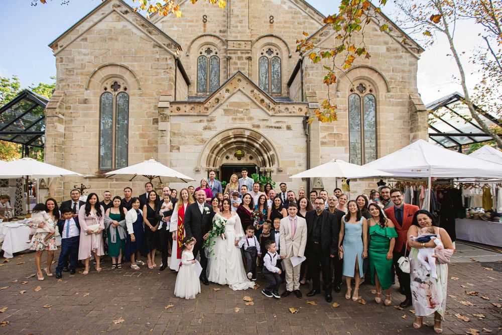 Large wedding party poses for group photo in front of stone church facade.