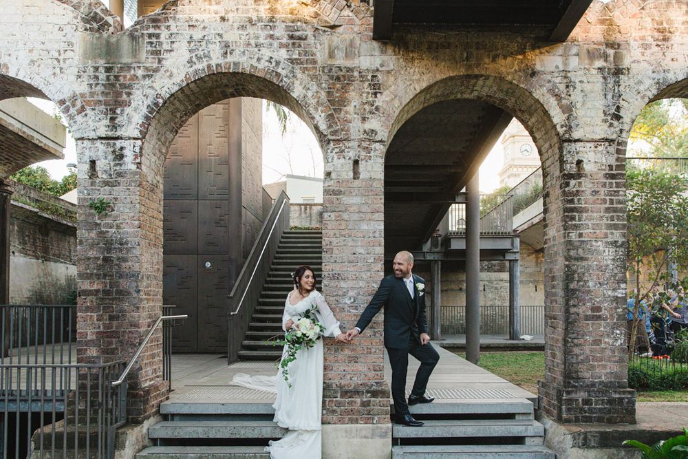 Wedding couple poses under brick archways and staircases in historic venue.