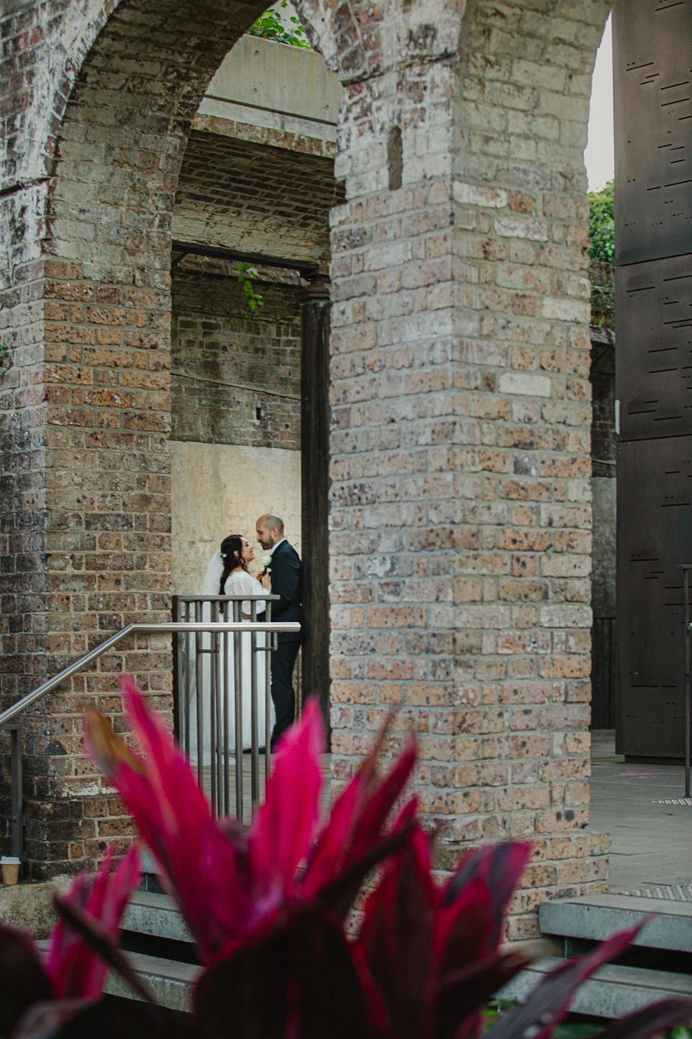 Romantic wedding moment captured under historic brick archways with vibrant pink flowers in foreground.
