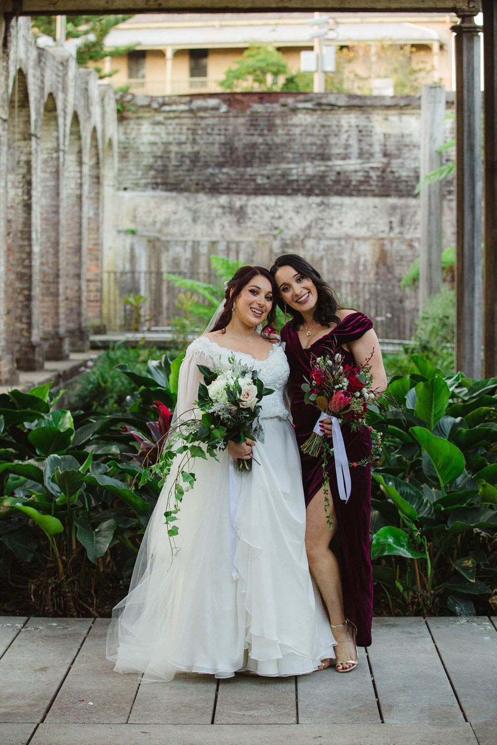 Wedding party in white dresses and burgundy attire pose along brick colonnade with lush greenery.