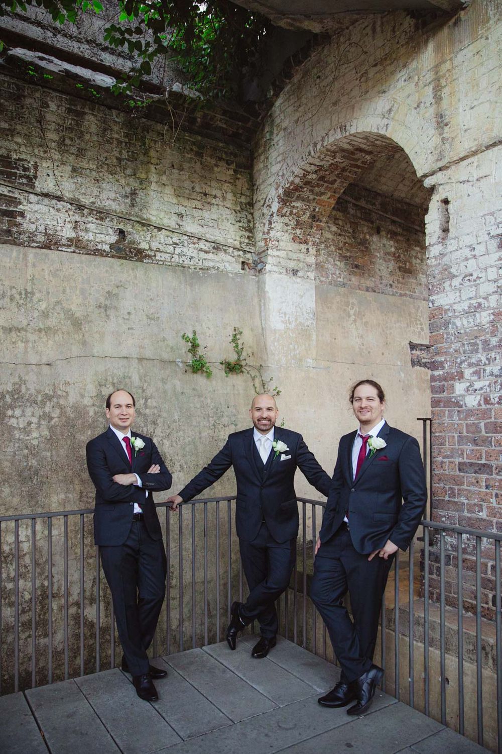 Groomsmen in black suits pose playfully against rustic brick wall and iron railing.