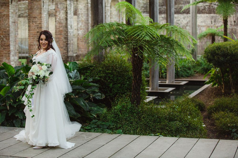 Wedding party members stand elegantly against weathered brick wall with green foliage.