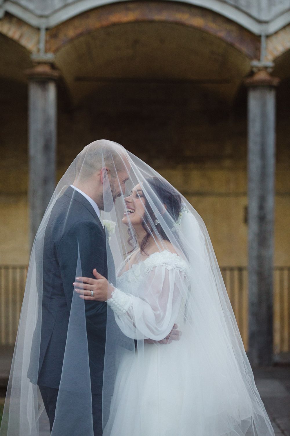 Intimate wedding moment captured under stone arches with flowing white veil.