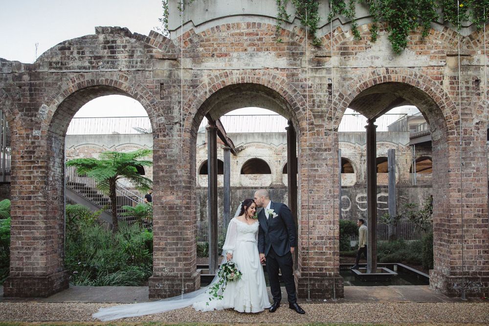 Wedding couple poses beneath series of rustic brick arches with greenery.
