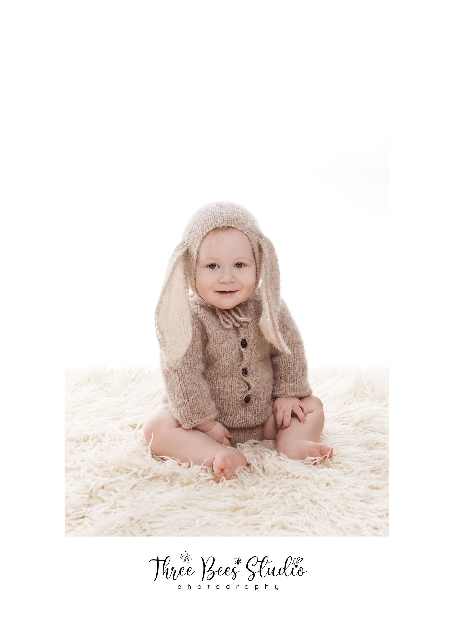 Baby wearing bunny-eared sweater sits on fluffy white rug in studio portrait.