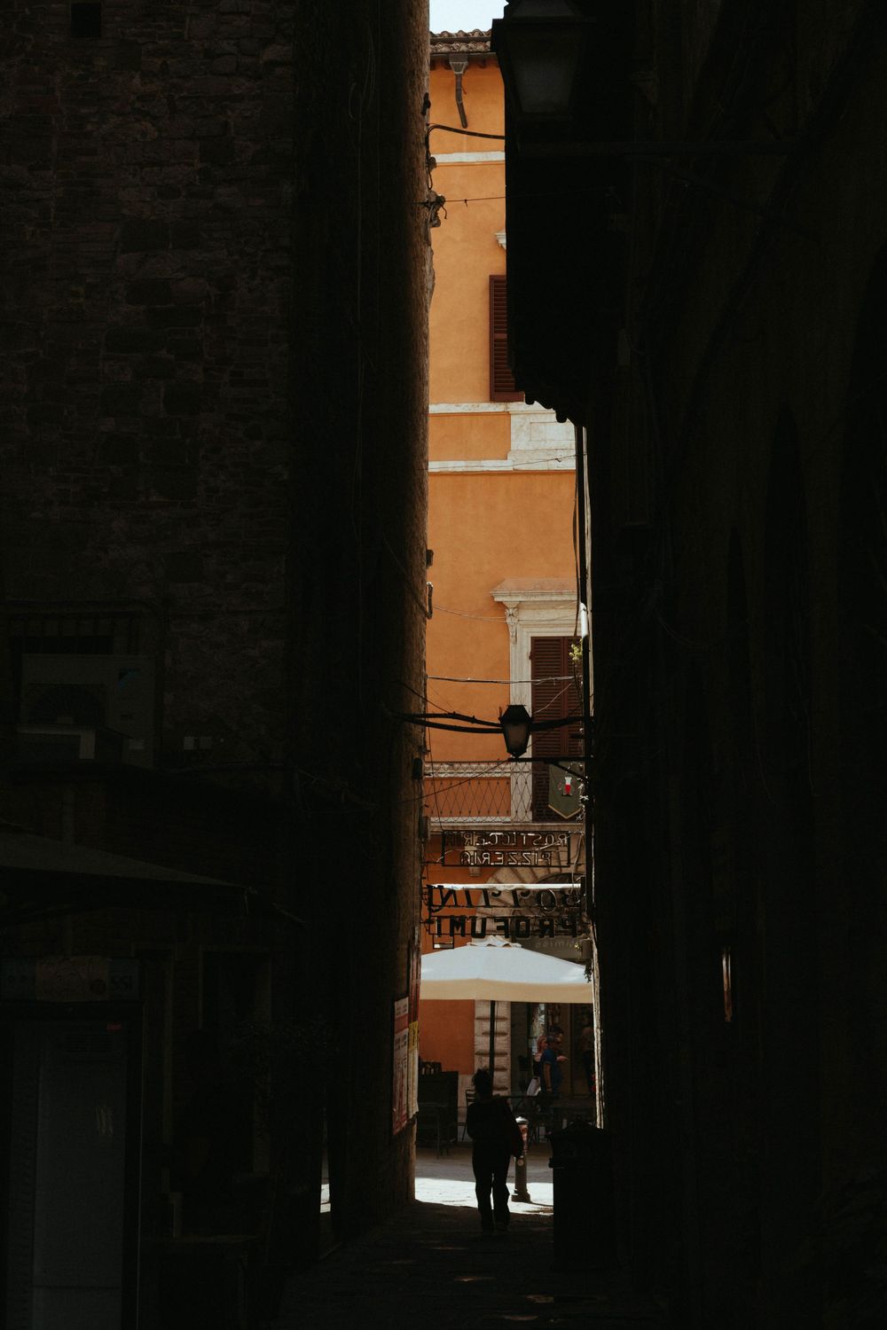 Narrow dark alley between buildings frames a sun-lit orange wall in an Italian town.