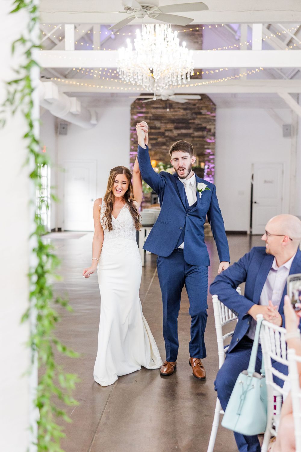 A newlywed couple makes their grand entrance into an elegant reception venue with white decor and crystal chandeliers.