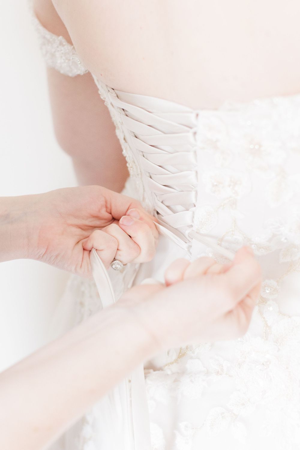 Close up view of hands lacing up the back of an elegant white wedding dress with delicate details.