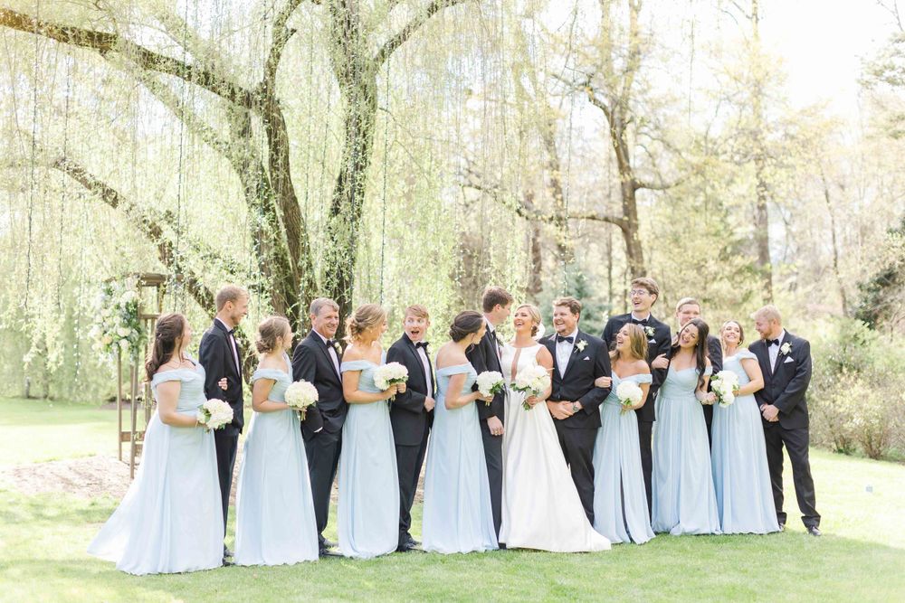 Wedding party in light blue dresses and black tuxedos posing under weeping willow trees.
