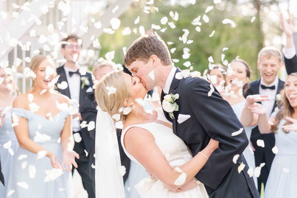 A wedding couple sharing a celebratory kiss while white flower petals fall around them.