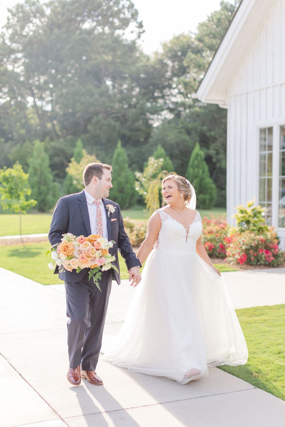 Joyful wedding couple walking outside white chapel with peach bouquet.