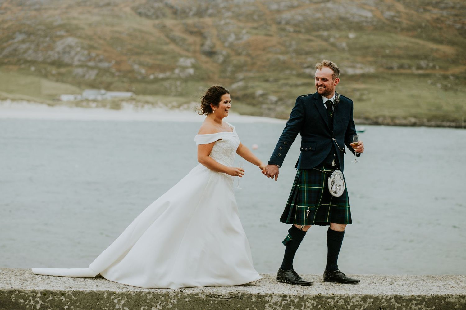 A bride in a white gown and groom in traditional Scottish kilt walking along a scenic mountain path.