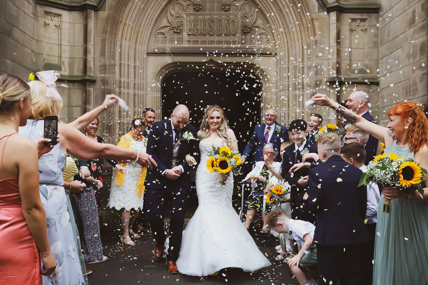 Wedding guests celebrating and throwing confetti outside a grand stone church entrance.