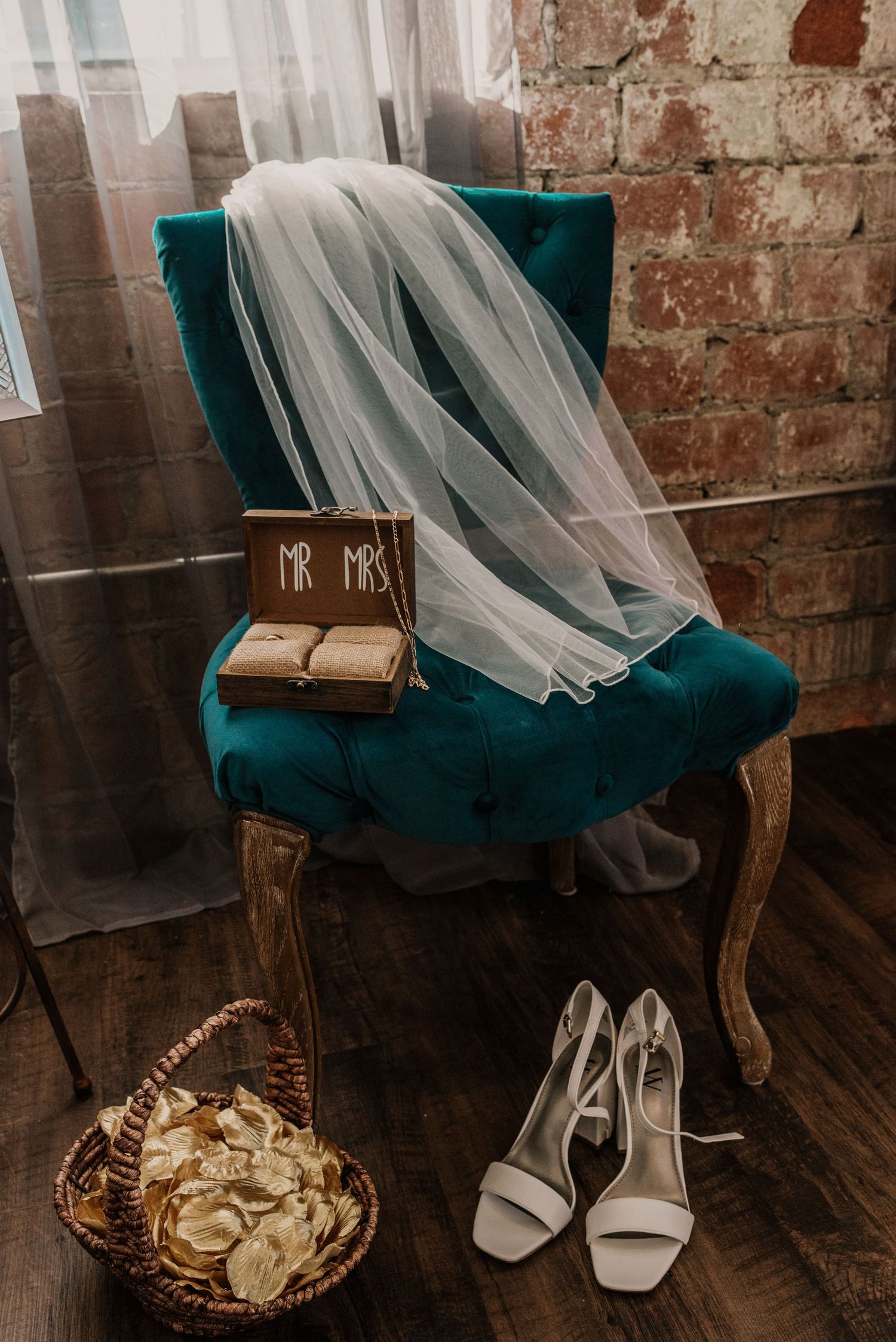 Wedding accessories displayed on teal velvet chair including veil, shoes and rings.