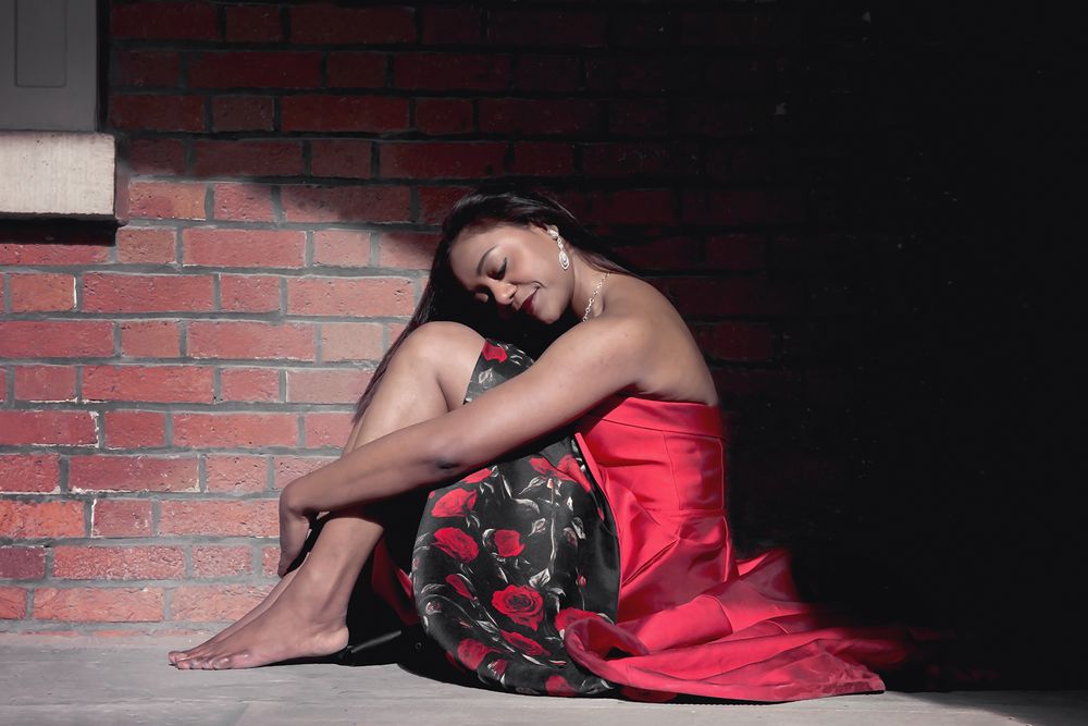 Person in red dress sitting against brick wall in dramatic lighting and dark setting.