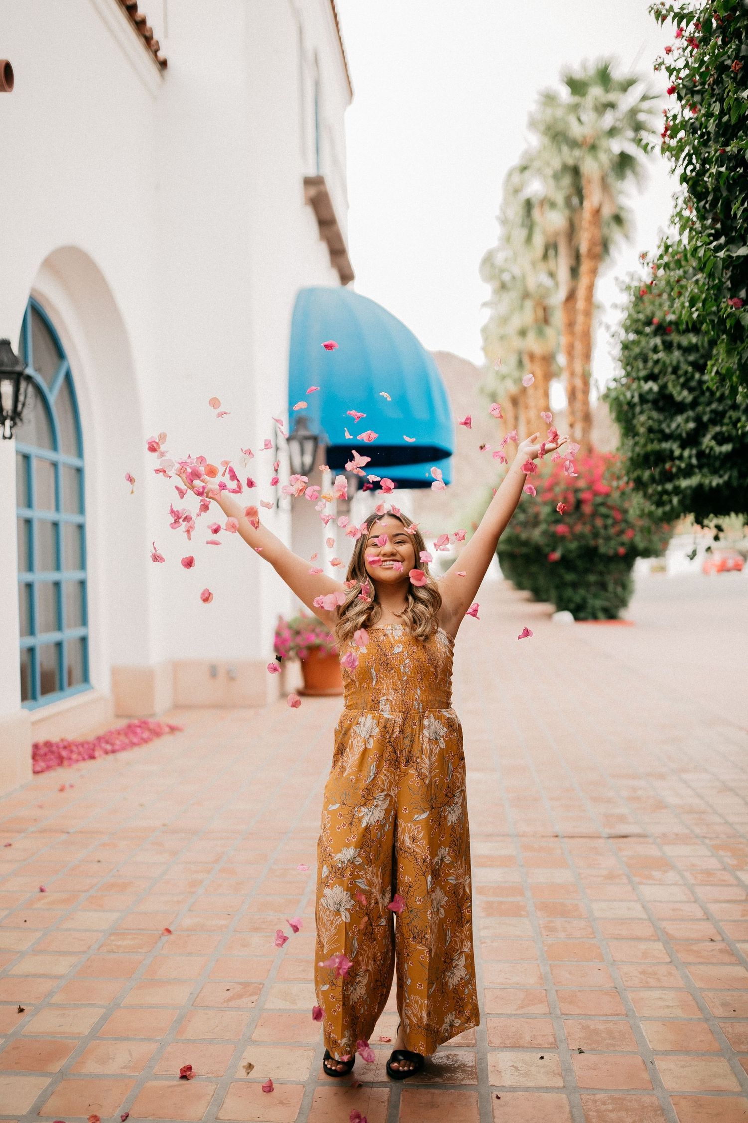 Person in floral jumpsuit joyfully throws pink flower petals in front of white Spanish-style building.