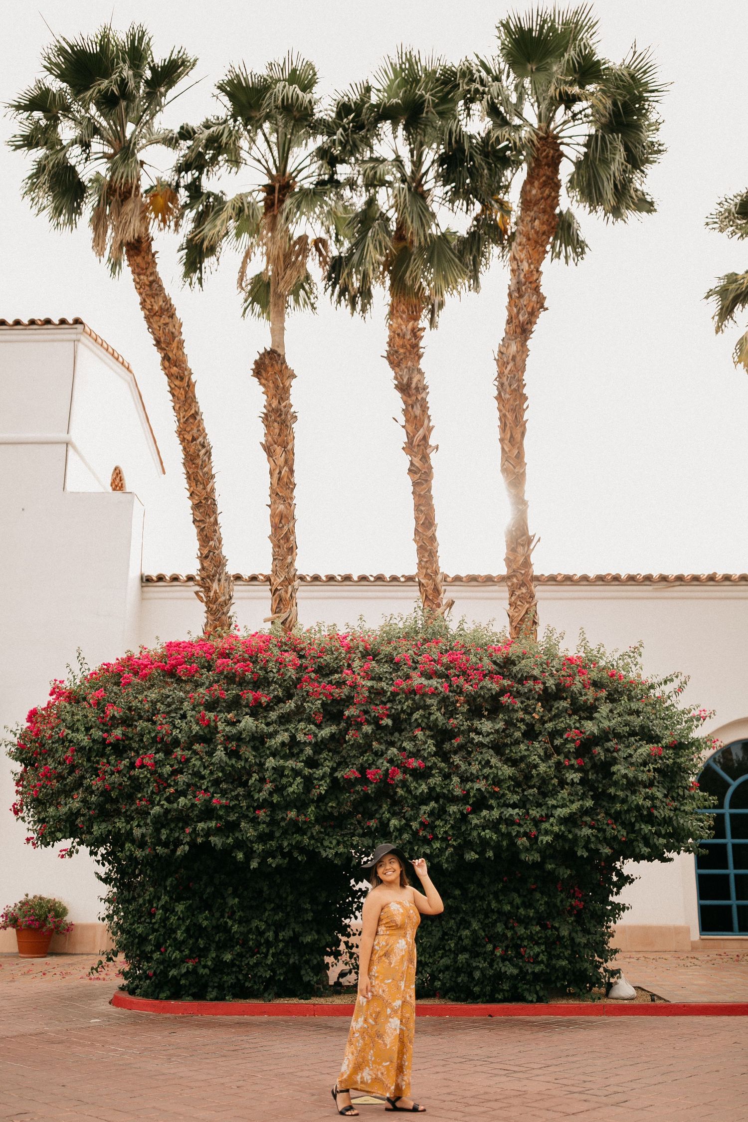 Person in yellow dress poses beneath tall palm trees and flowering bushes at Spanish-style building.