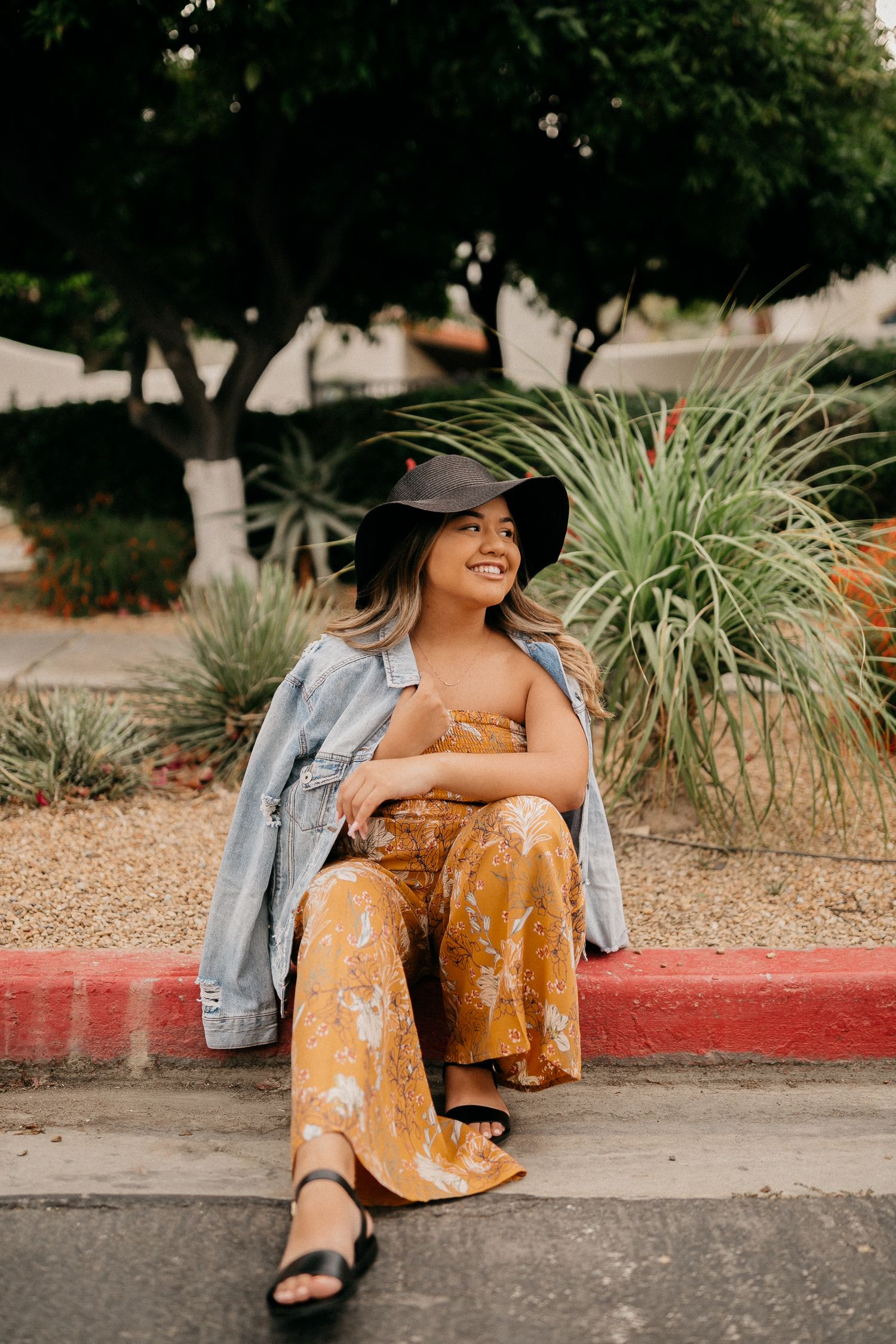 Casual portrait of person in denim jacket and yellow jumpsuit sitting on curb with tropical plants.
