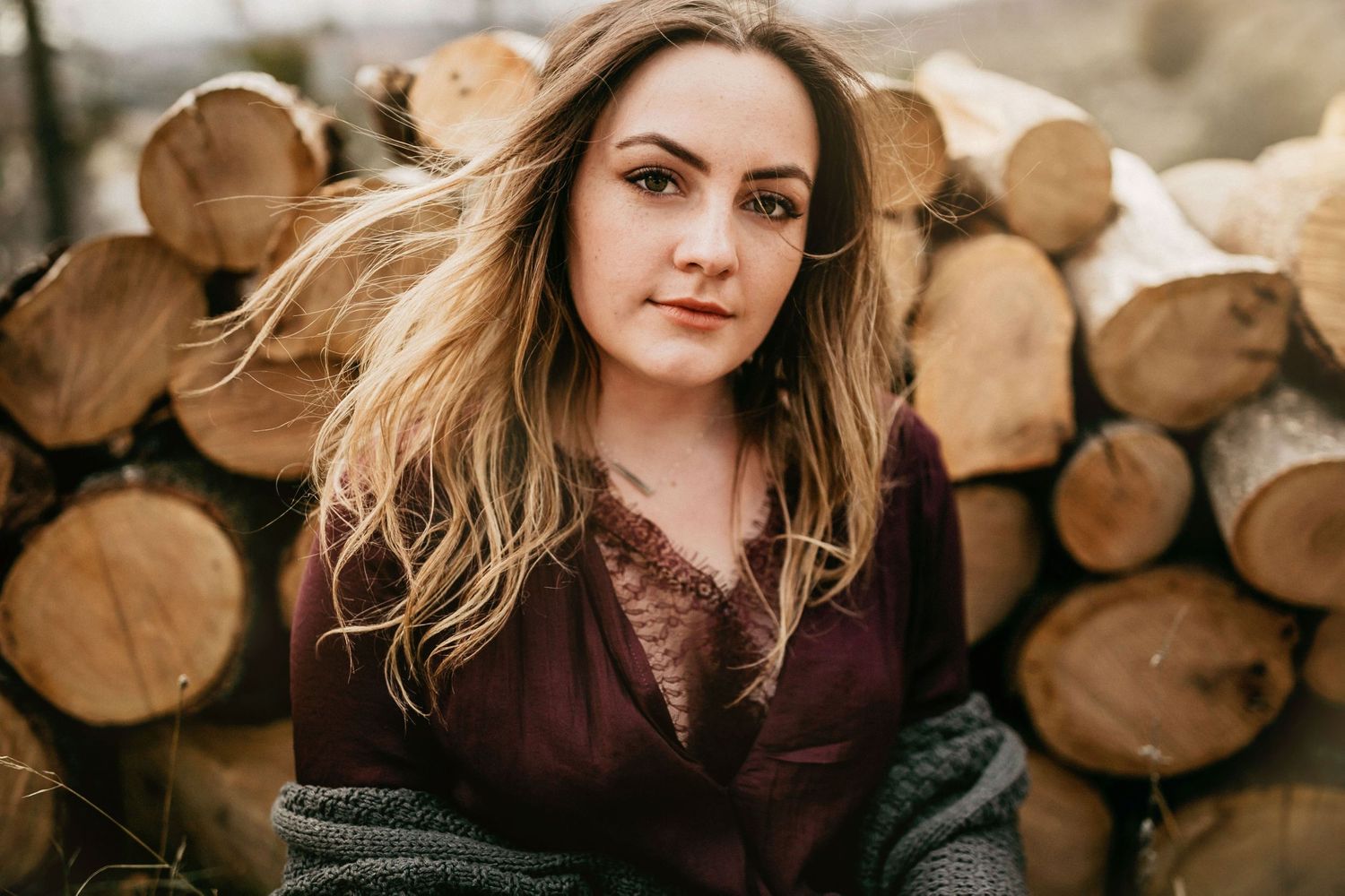 Outdoor portrait with woodpile background wearing burgundy sweater.