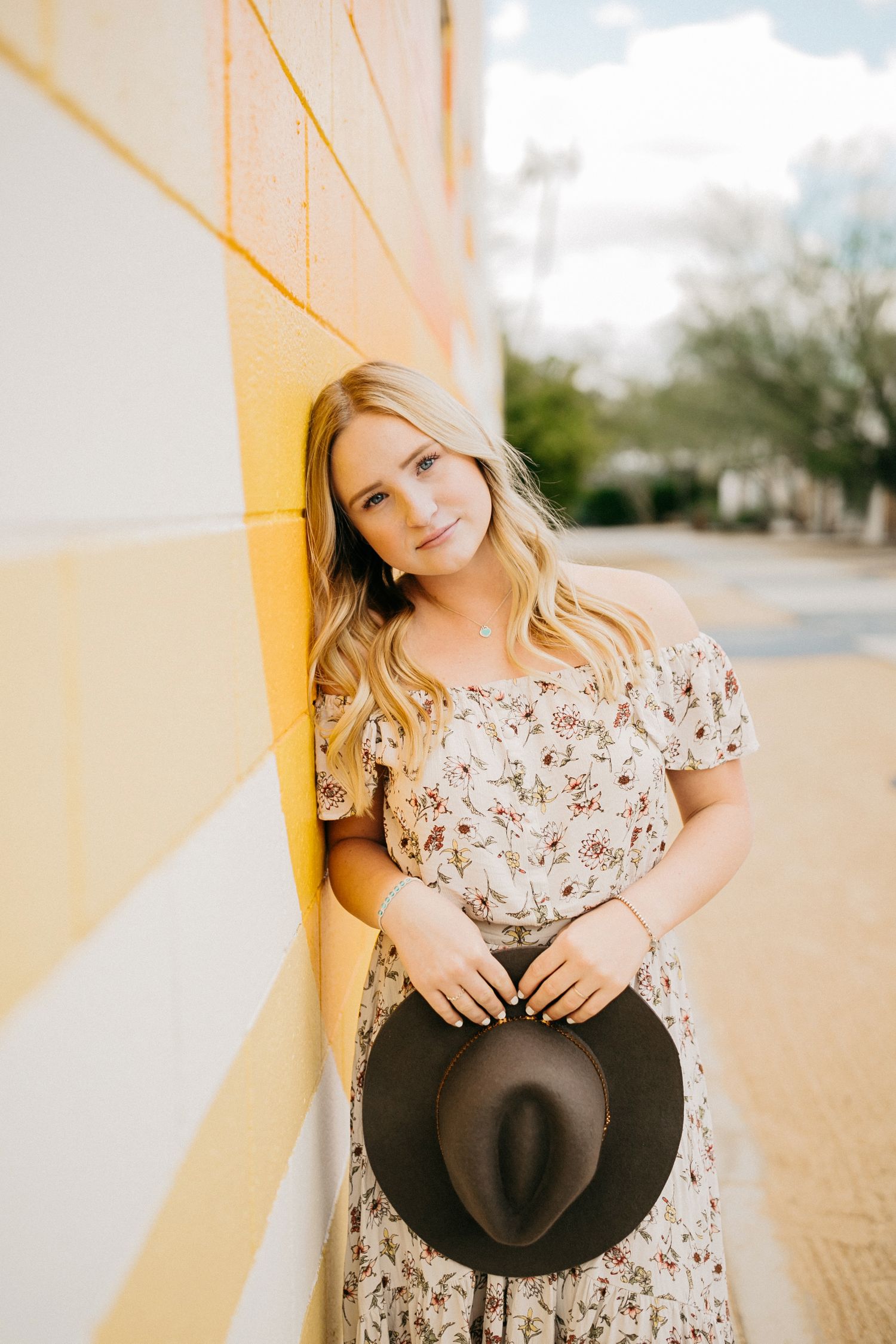 Portrait against yellow wall holding dark hat wearing floral dress.