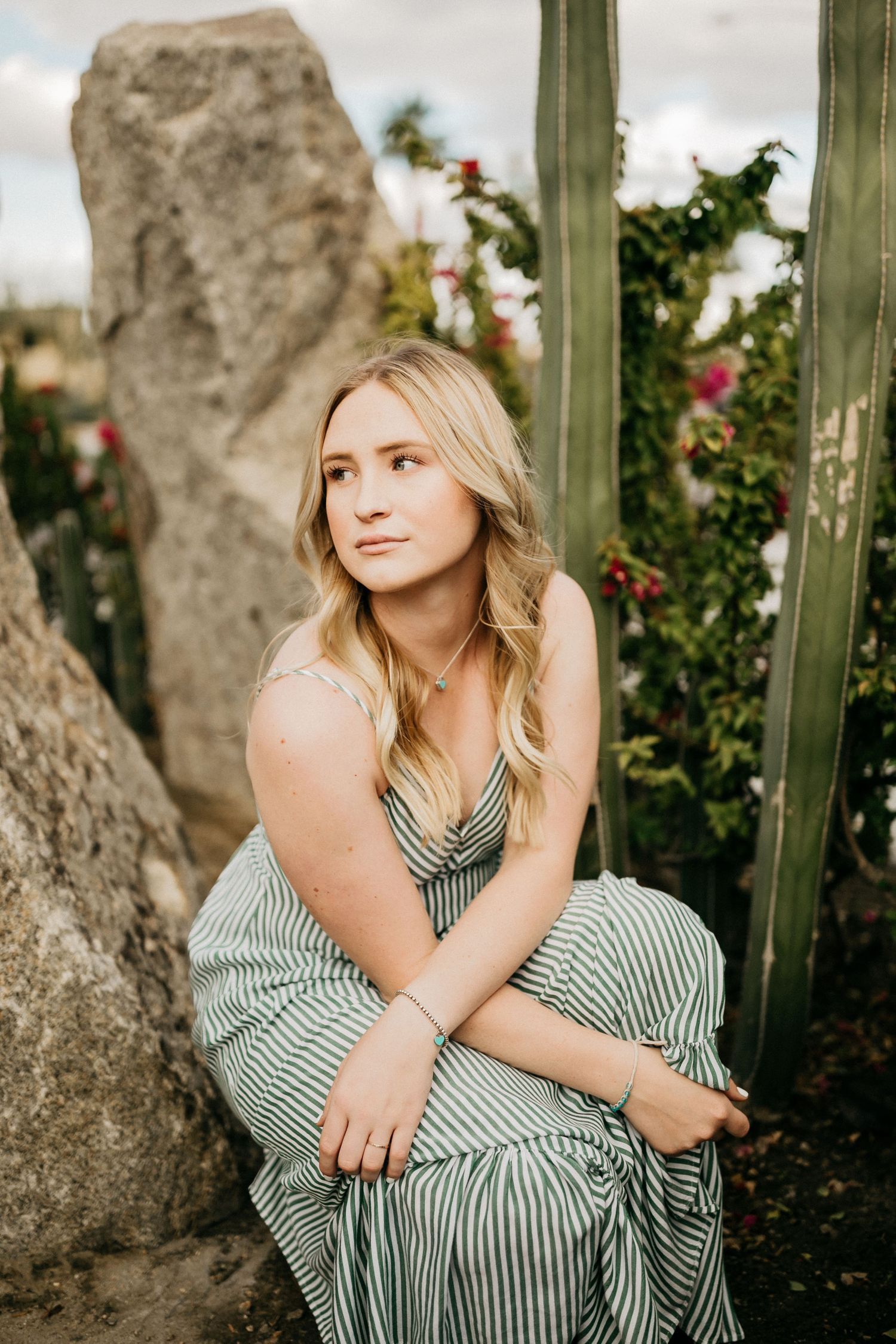 Portrait in striped dress seated among garden rocks and flowering plants.