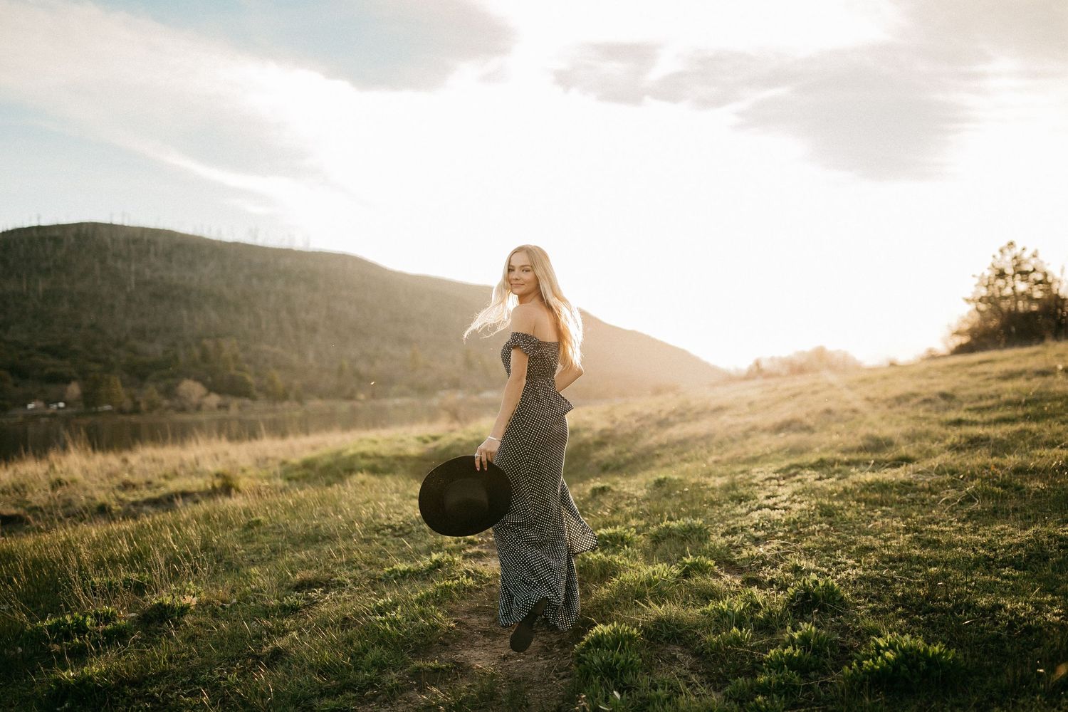 Figure silhouetted against sunset holding hat on grassy hilltop with mountains in background.