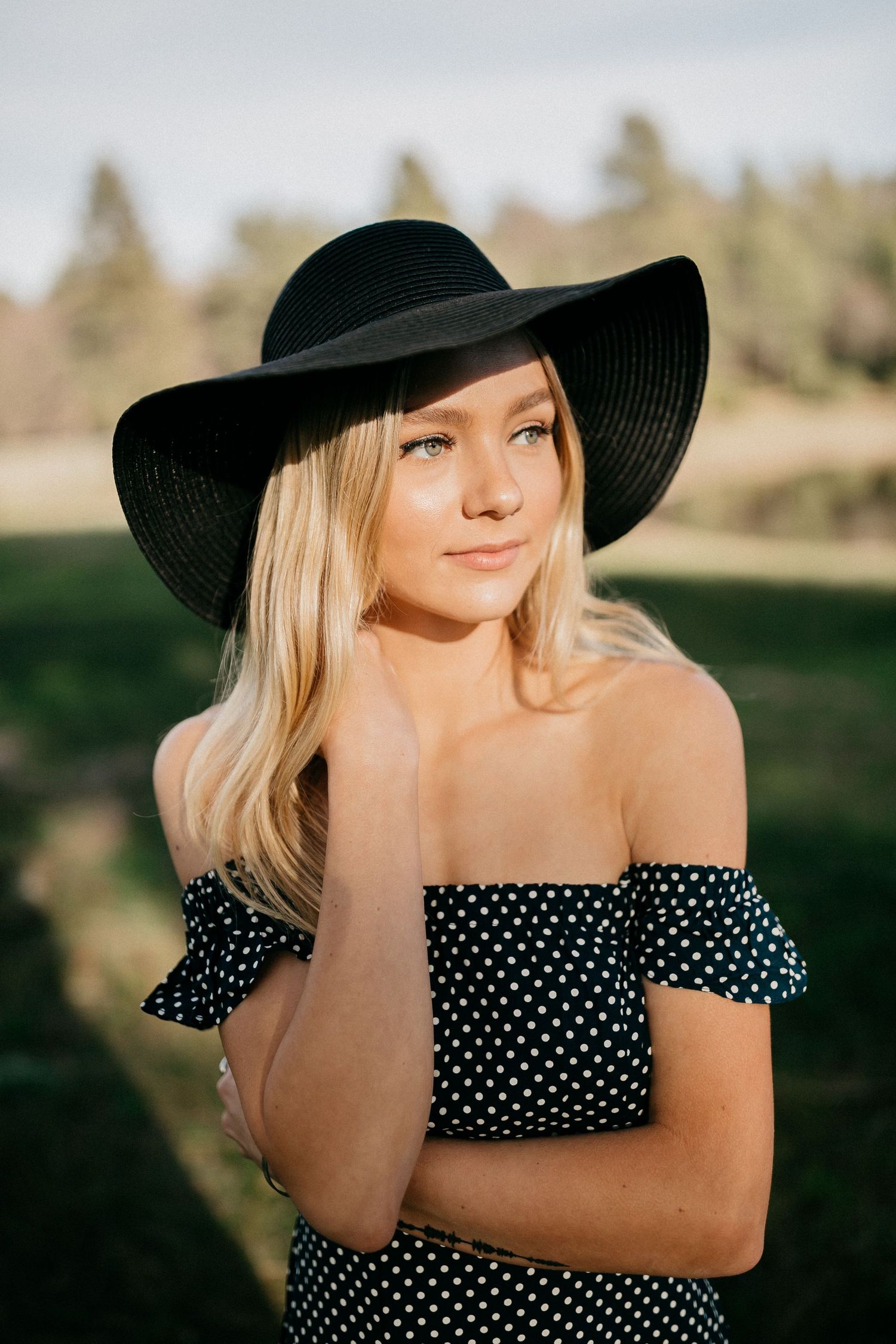 Woman in polka dot top and wide-brim black hat posing outdoors at sunset.