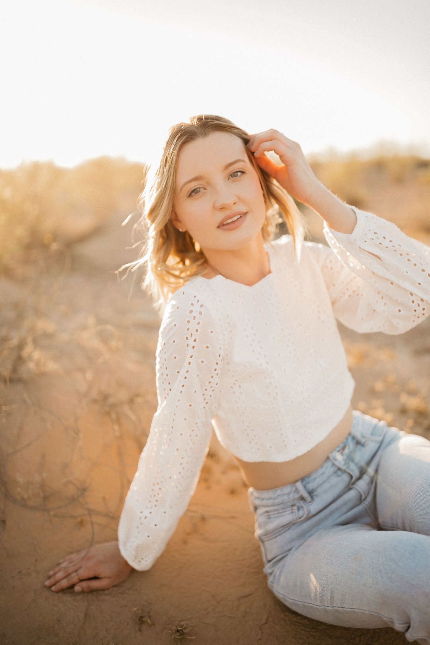 Sunset portrait in white eyelet top and jeans on sandy terrain.