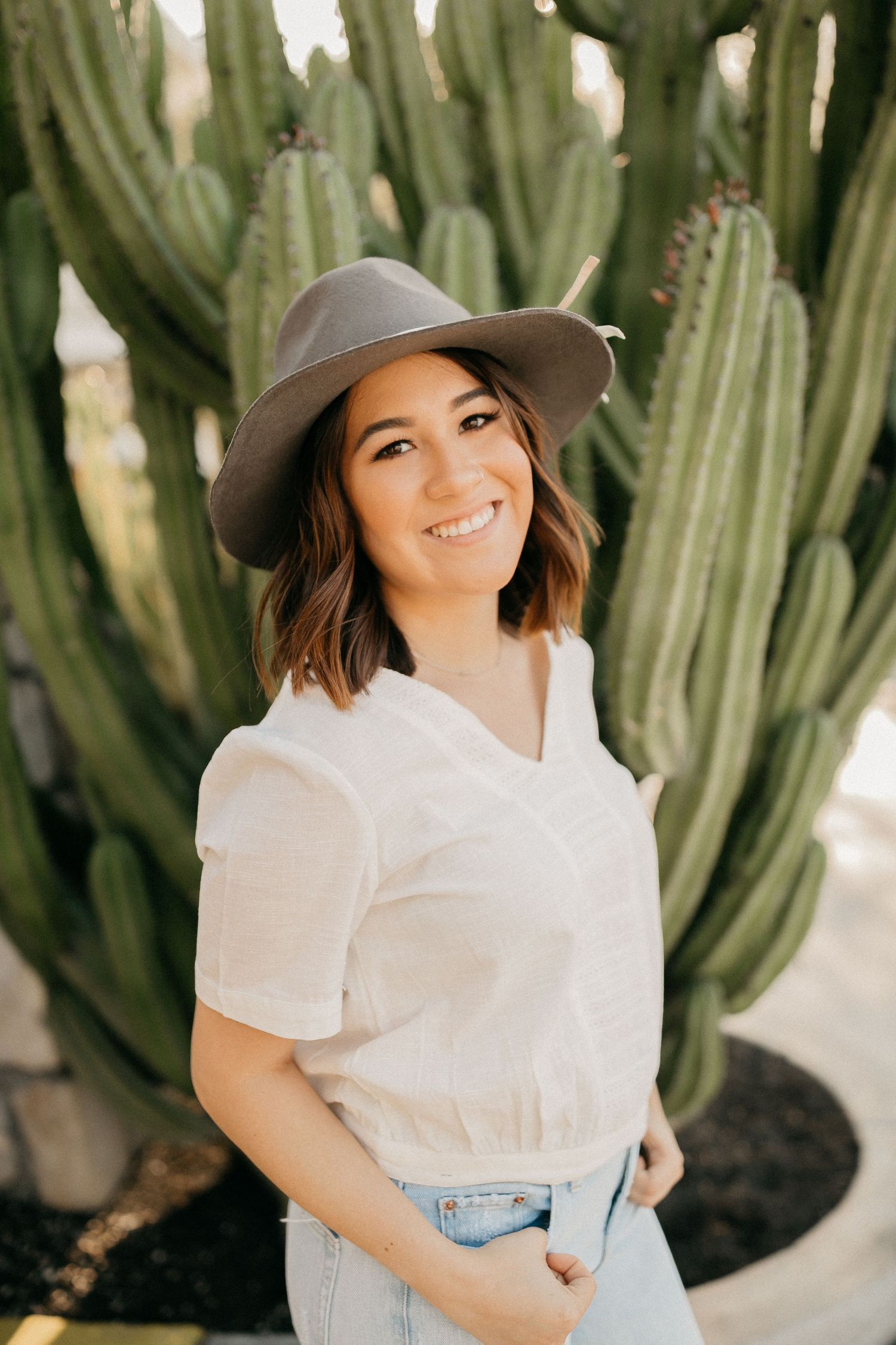 Woman in white shirt and gray hat smiling in front of tall desert cactus.