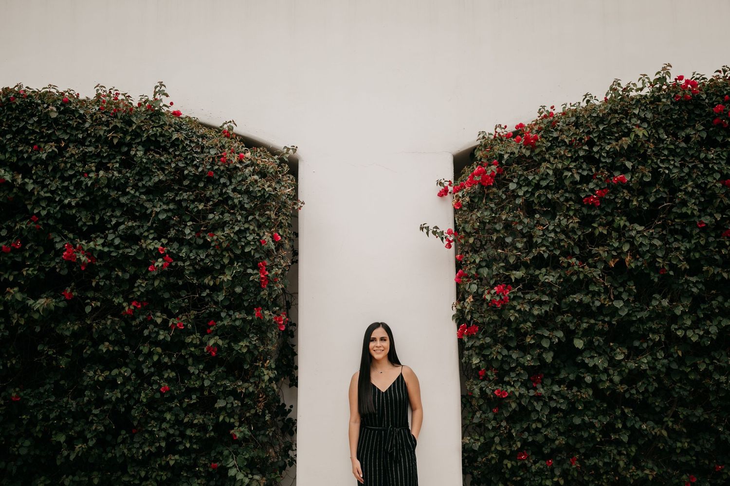 Dramatic portrait between green hedges with red flowers against white wall.