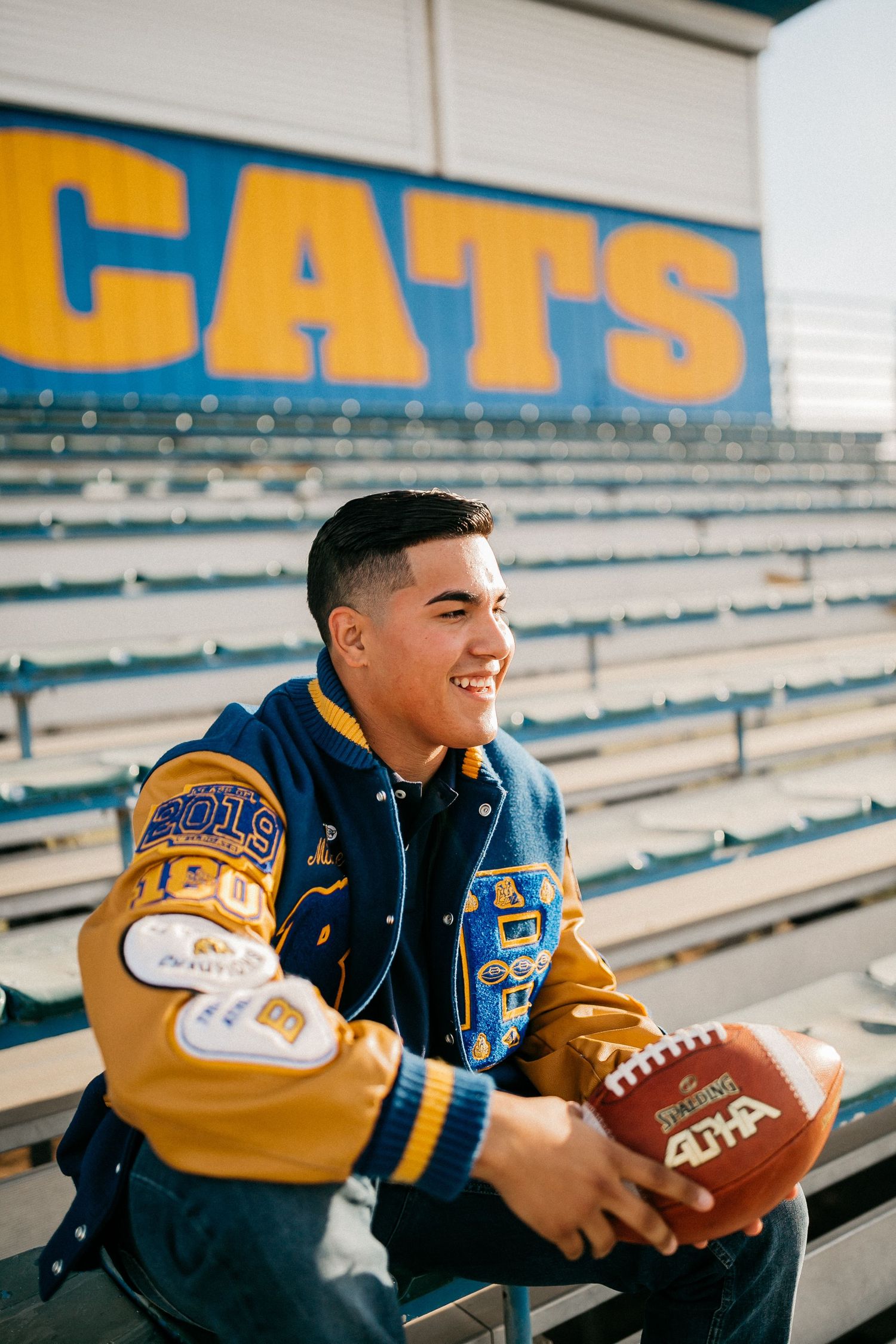 Student athlete in blue and gold varsity jacket sits in bleachers holding a football.