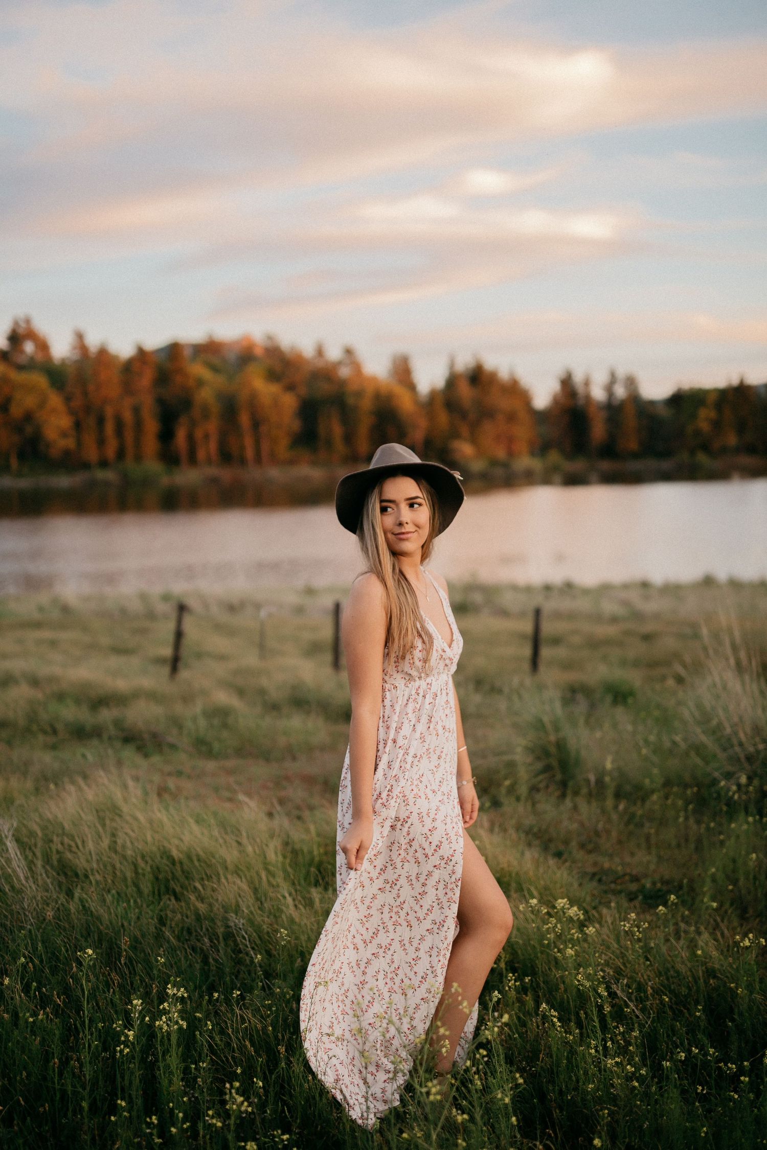 Person in floral maxi dress poses in field during golden hour with lake and autumn trees behind.