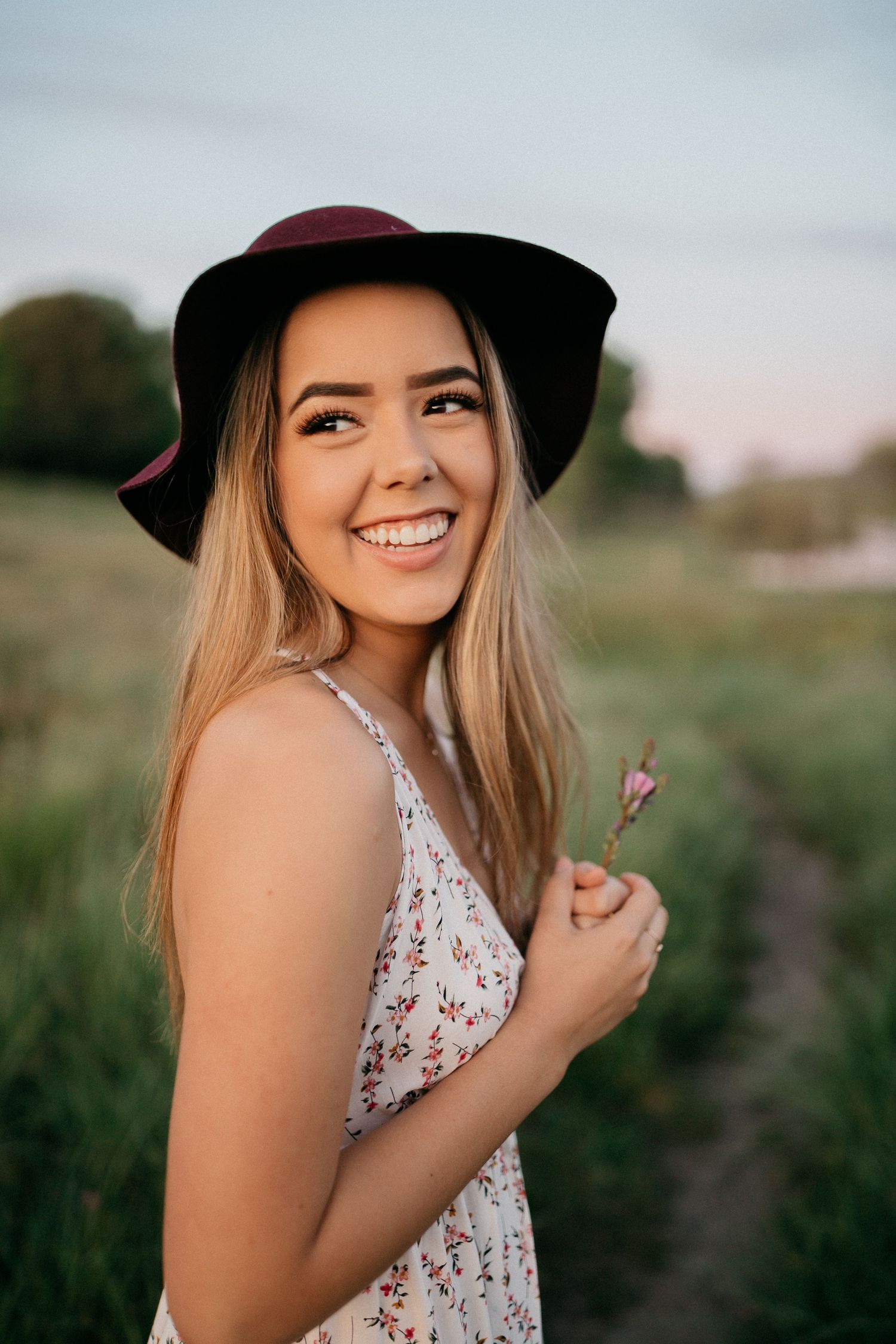 Portrait of someone in burgundy hat holding flower against natural outdoor backdrop.
