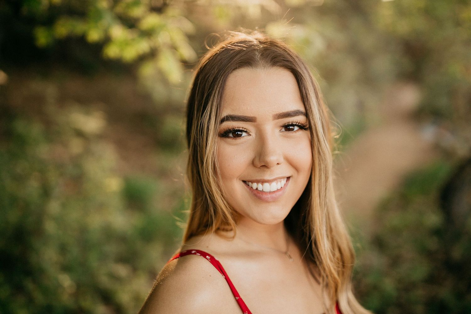 Portrait of a smiling woman with long brown hair wearing a red top against a blurred natural background.