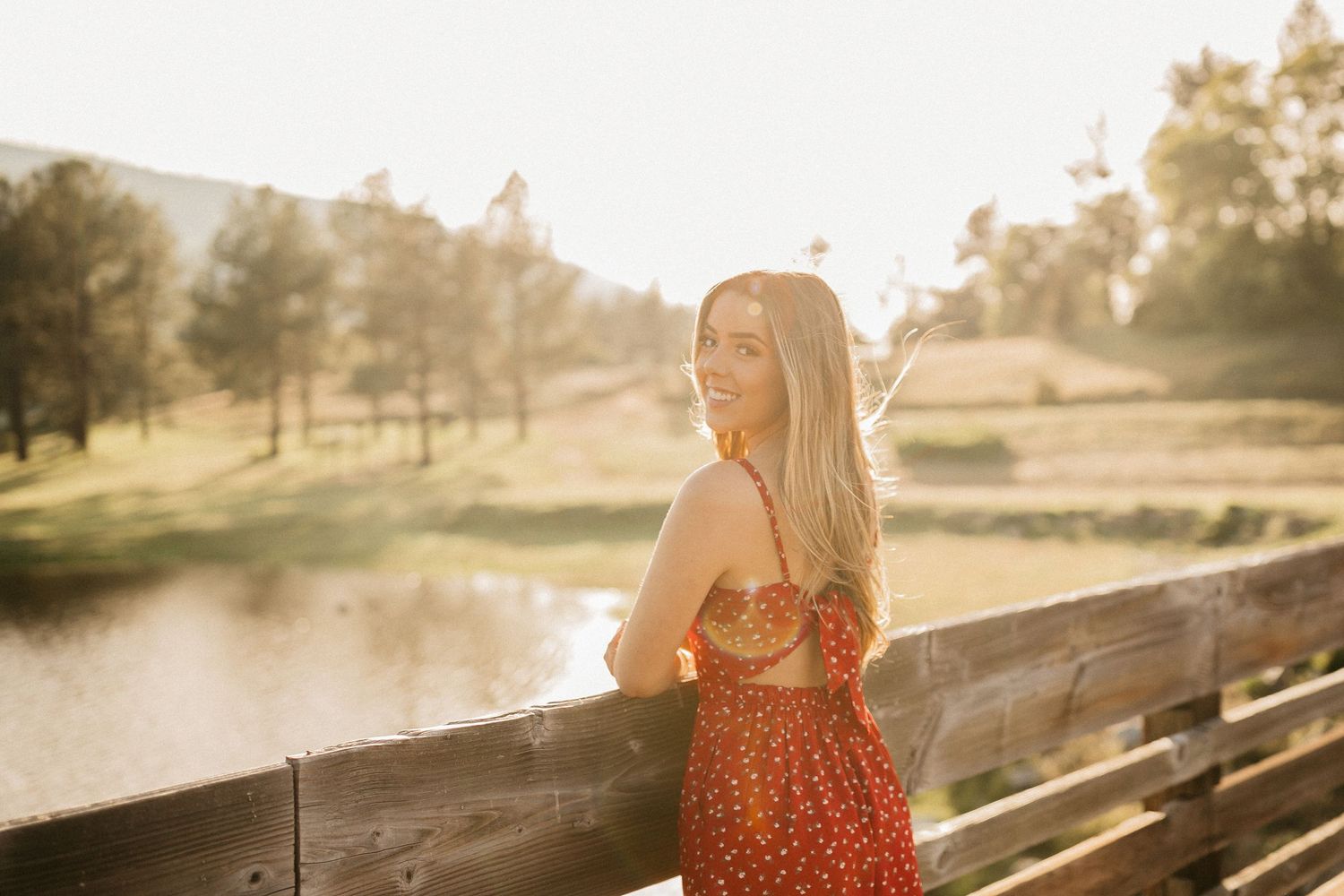 Golden sunlight illuminates figure in red dress standing on wooden bridge near pond.
