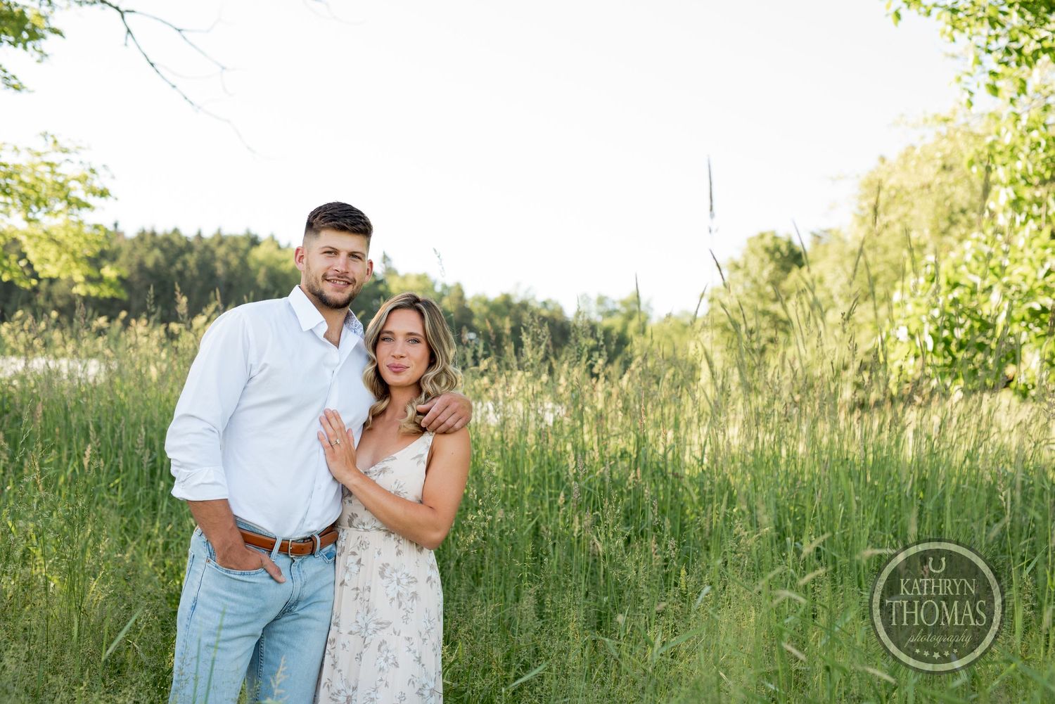 A romantic couple embraces in a sunlit meadow with tall grass and trees in the background.