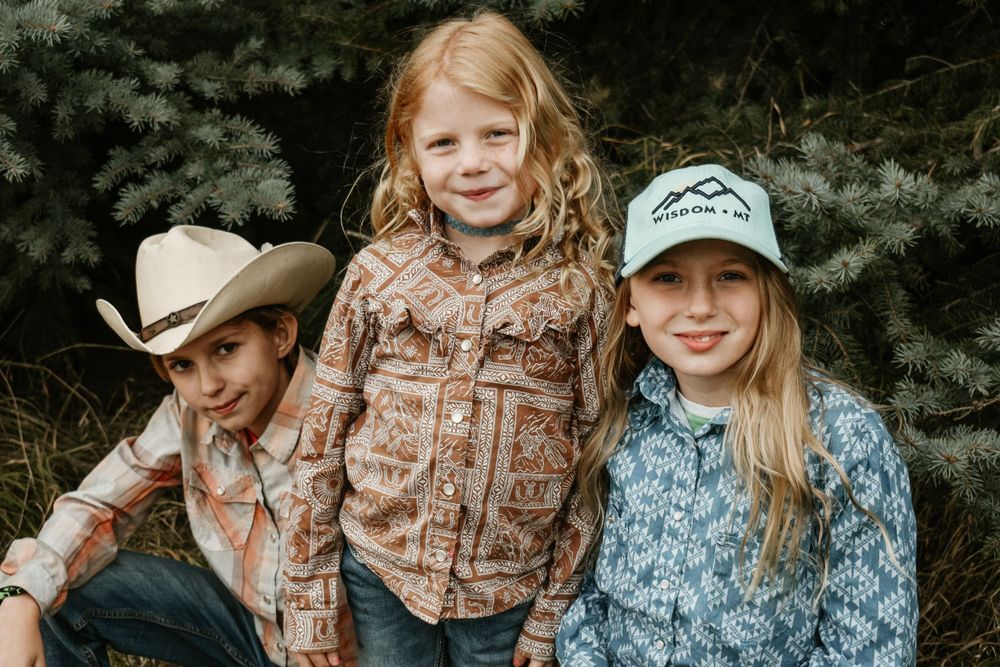 Three people in western wear pose together against a pine tree backdrop.