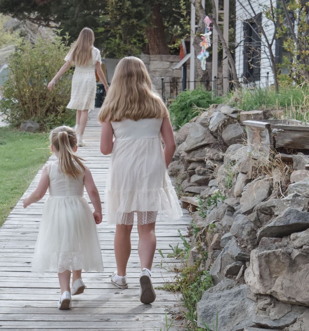 Three people in white dresses walk along a wooden boardwalk near stone walls.