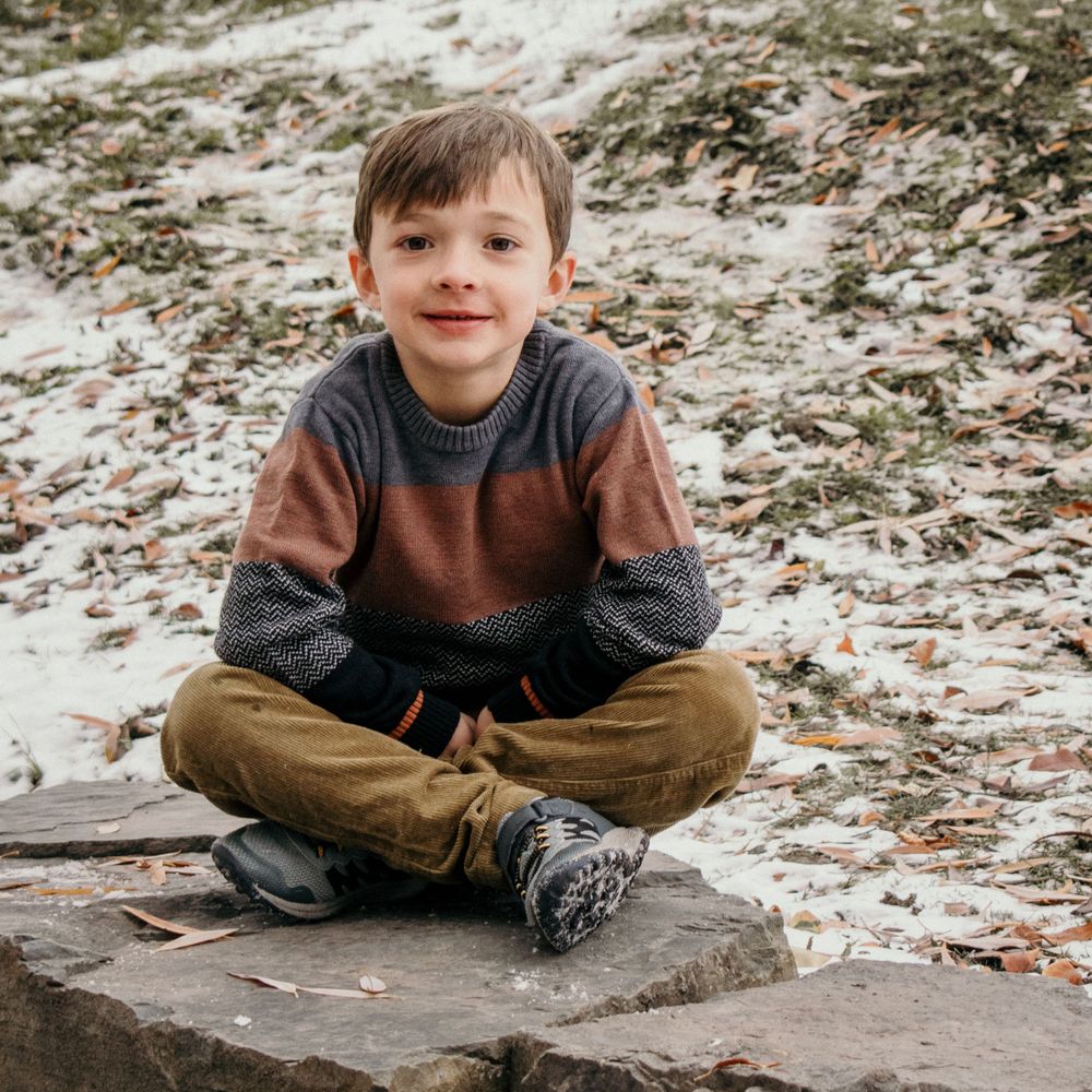 Young child sitting cross-legged on a stone surface covered in snow.
little boy, family photography