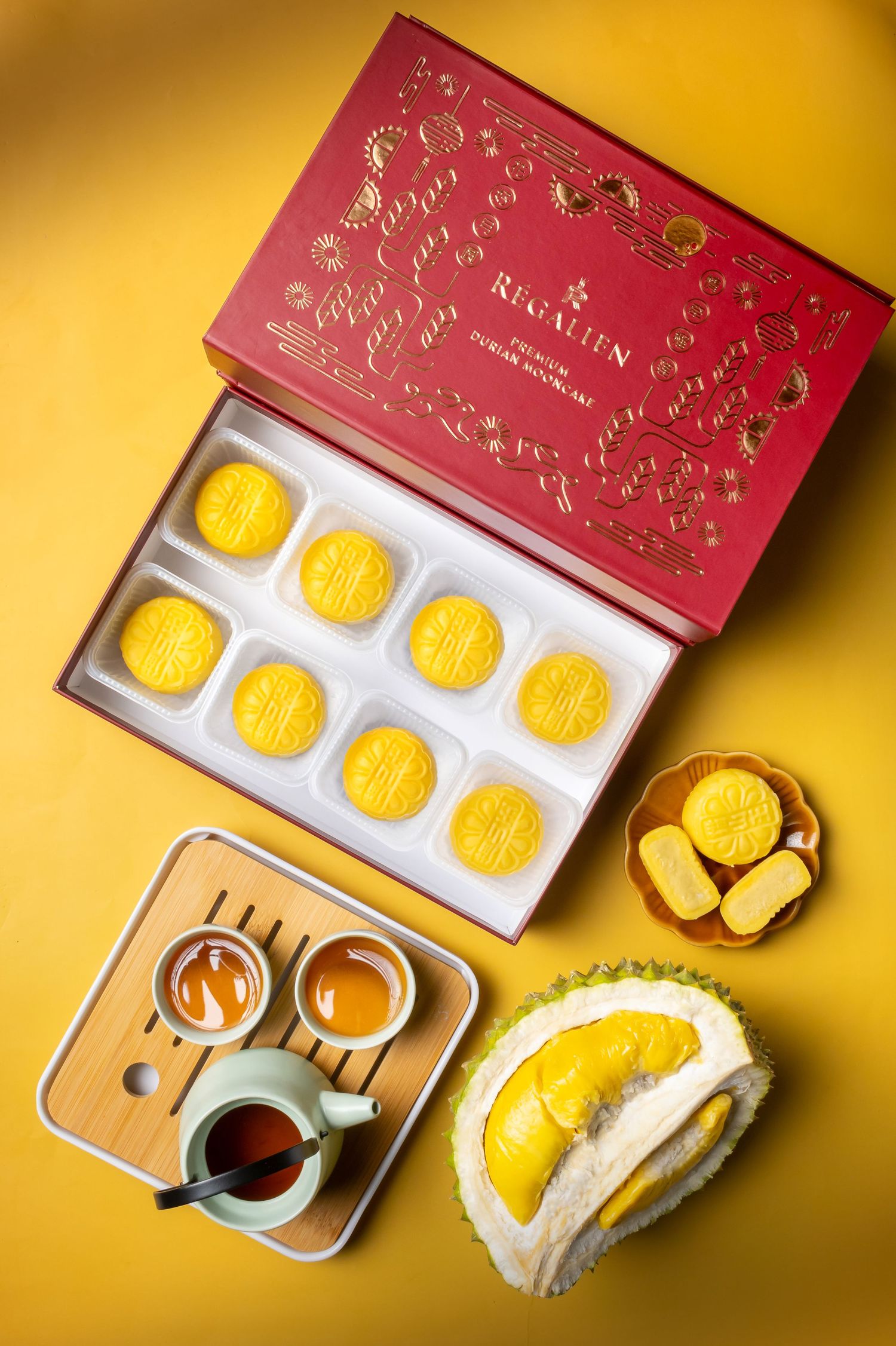 Elegant red gift box containing yellow mooncakes with tea and durian dessert on golden background.