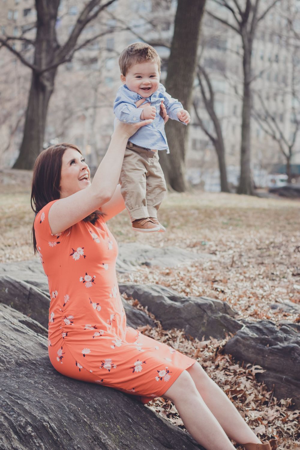 Woman in coral dress playing with child on fallen log in autumn park.