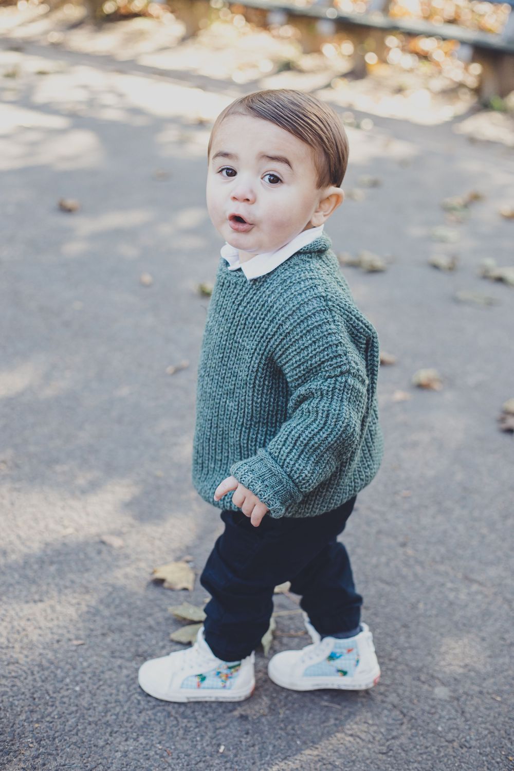 Toddler in green sweater and sneakers walking on pavement.
