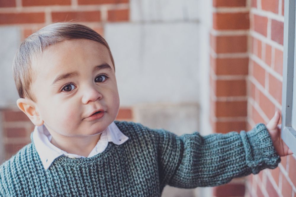 Child in grey knit sweater smiling indoors near brick wall.