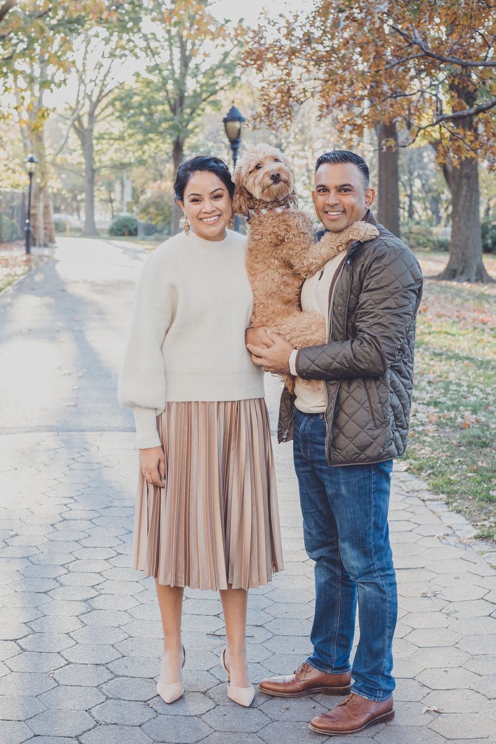 A couple poses with their small dog on a stone path surrounded by autumn trees.