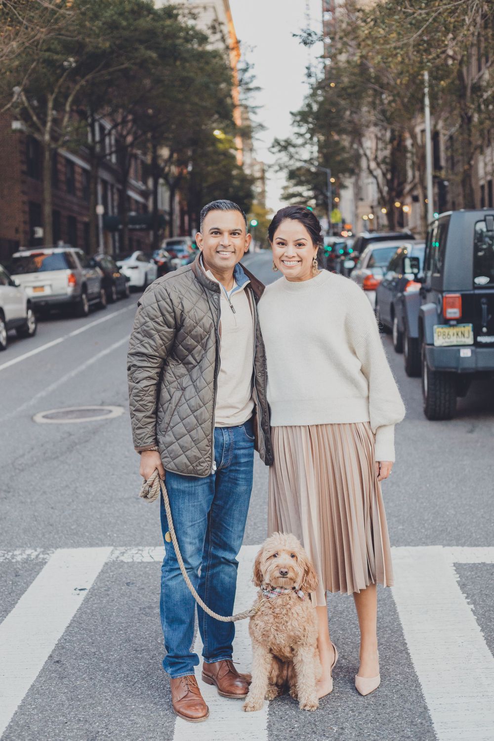A couple walks their small dog across an urban crosswalk on a city street.