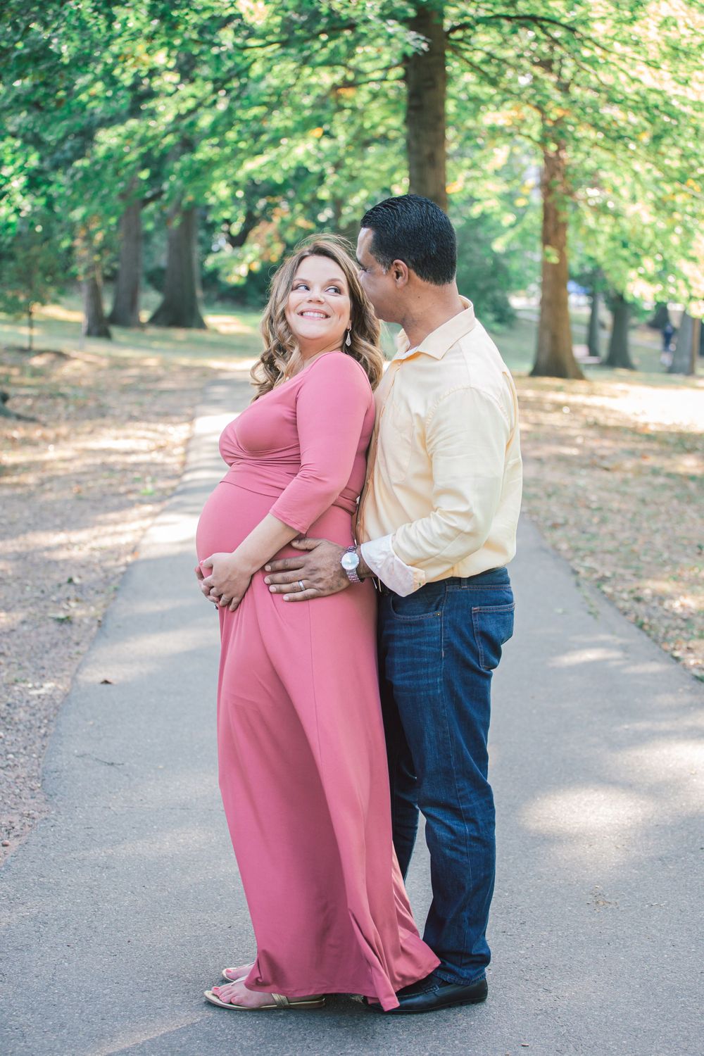 A couple shares an intimate moment on a tree-lined path, wearing pink dress and cream shirt.