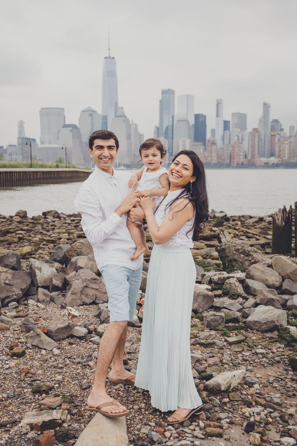 Family poses on rocky shore with Manhattan skyline visible in background.