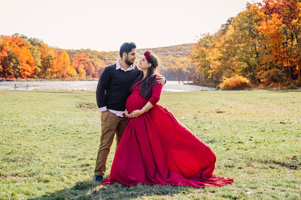 Couple embracing in field with vibrant autumn foliage and red dress flowing in wind.